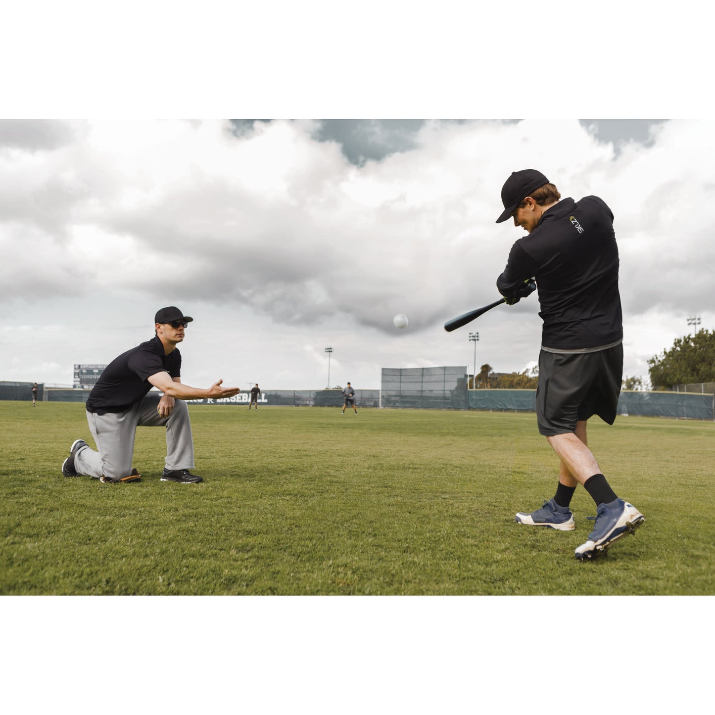 Person practicing baseball swing while coach crouches on a grassy field outdoors