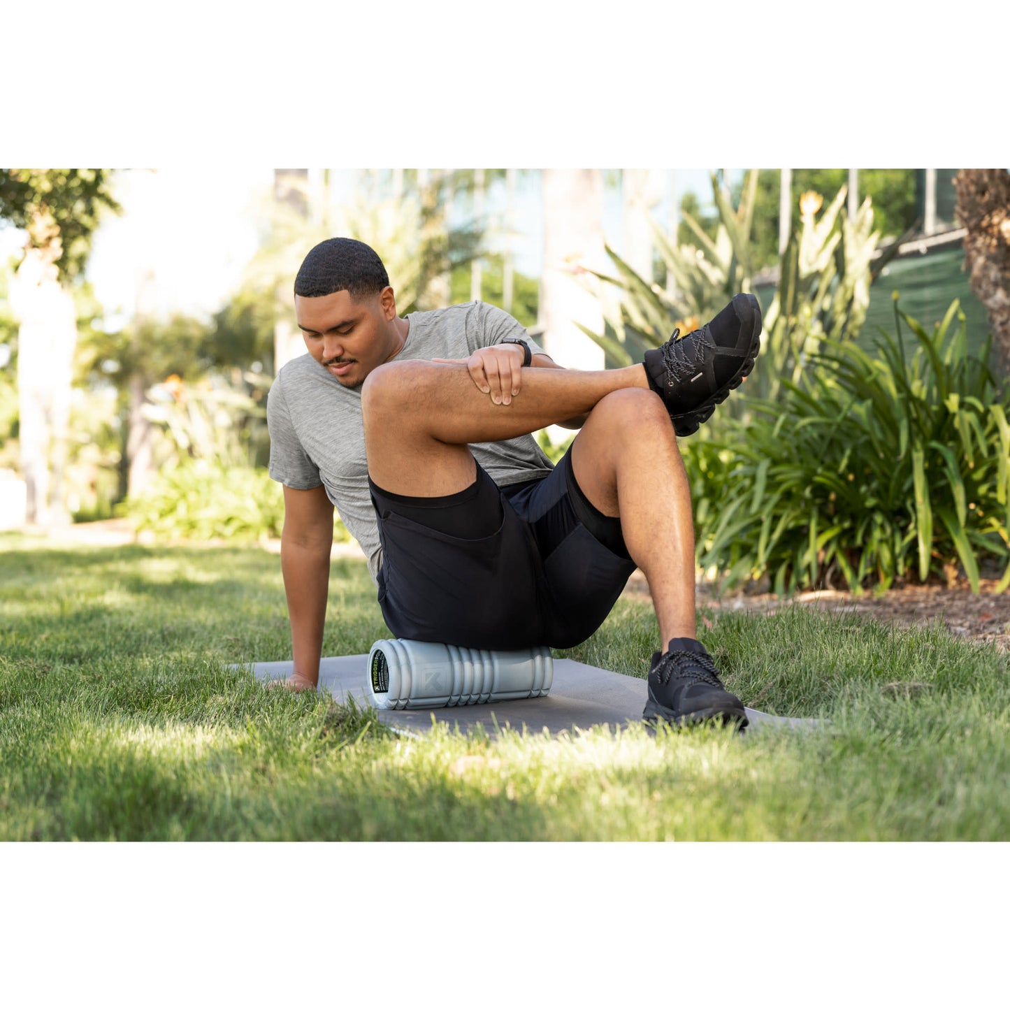 Man stretches leg using foam roller on grass in a park during outdoor exercise.