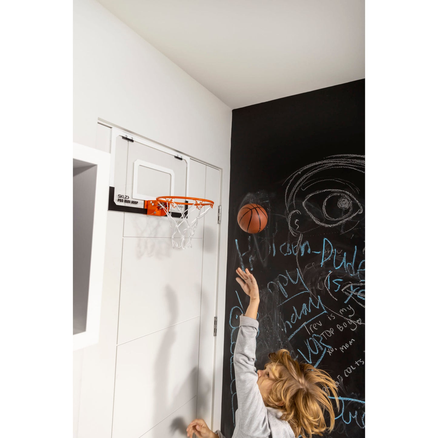 Child throws a basketball towards a hoop on a black chalkboard wall in a room.