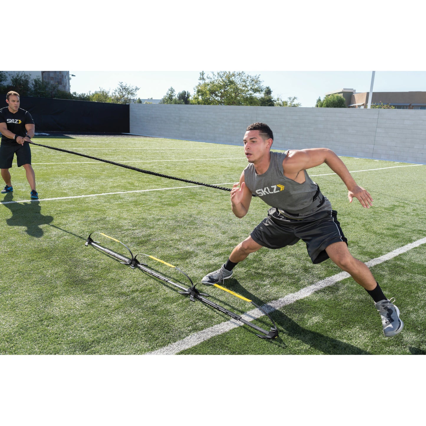 Athlete pulls resistance band during training on a grassy field with two trainers and a wall in background