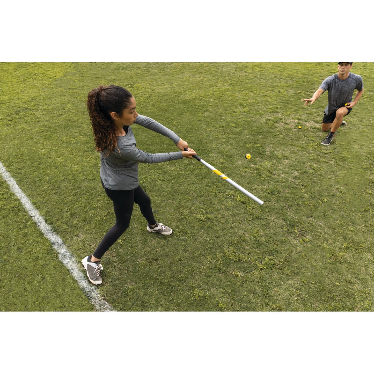 Girl swings a bat at a baseball on a grassy field with a man kneeling nearby