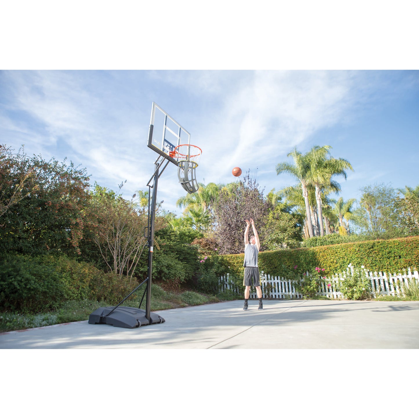 Person shoots basketball on outdoor driveway with trees and blue sky in the background