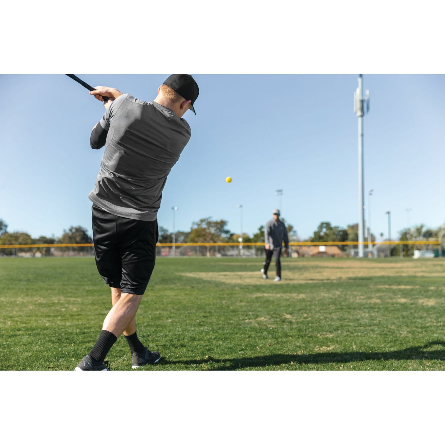 Person swings golf club on a field with a yellow ball in mid-air under a clear sky