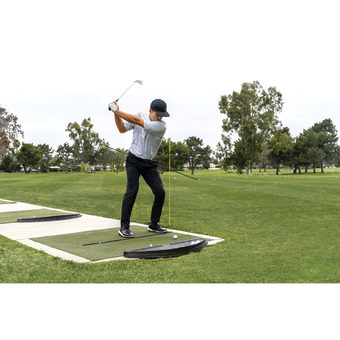 Golfer swings club on practice mat at outdoor driving range with trees and cloudy sky