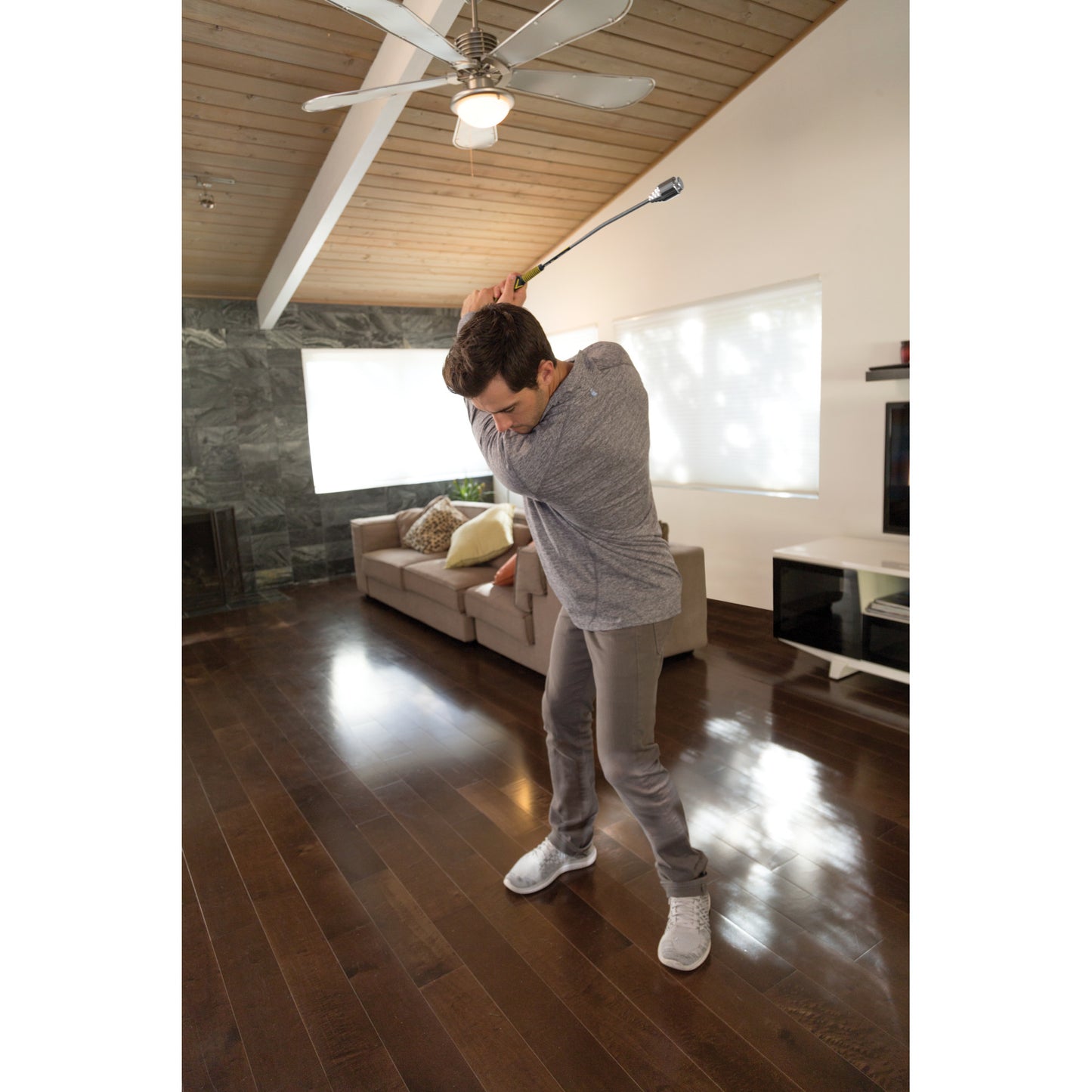 Man swings golf club indoors on wooden floor in living room with sofa and ceiling fan