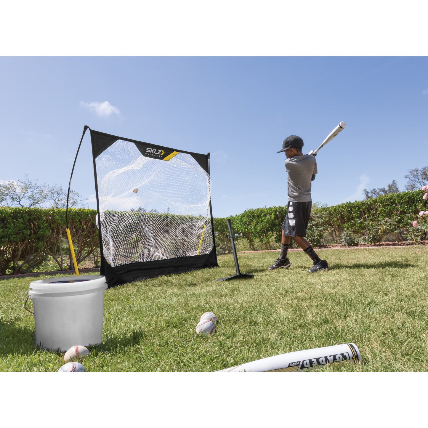 Boy practices baseball hitting near a net with a ball, surrounded by grass and shrubs.