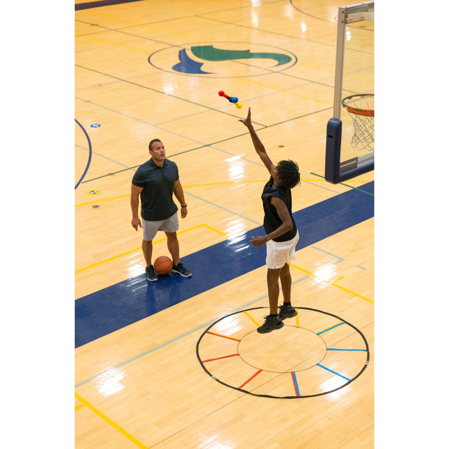 Player shoots basketball near coach in gymnasium with a colored circle on floor