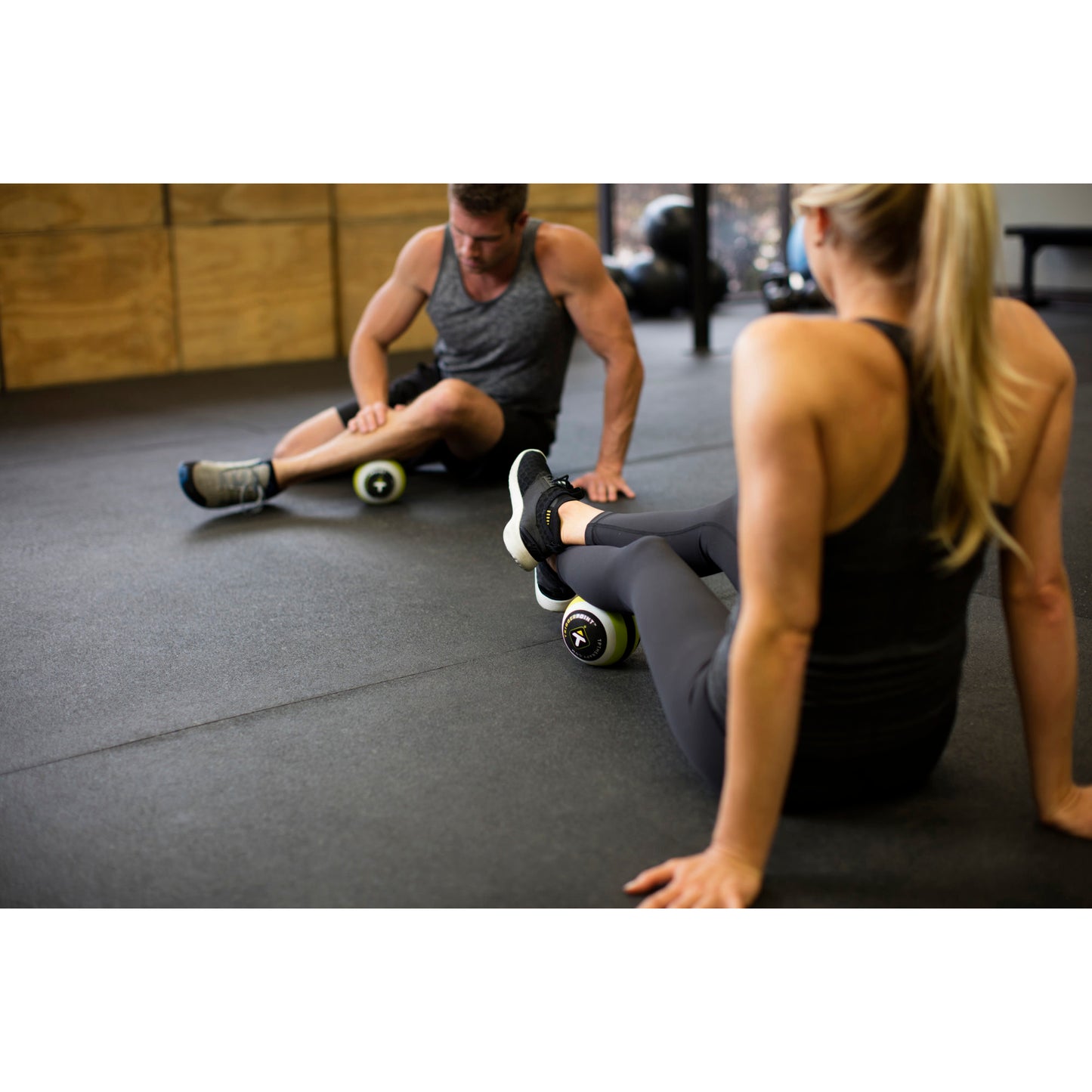 A man and a woman use massage rollers on their legs during a workout session in a gym environment.