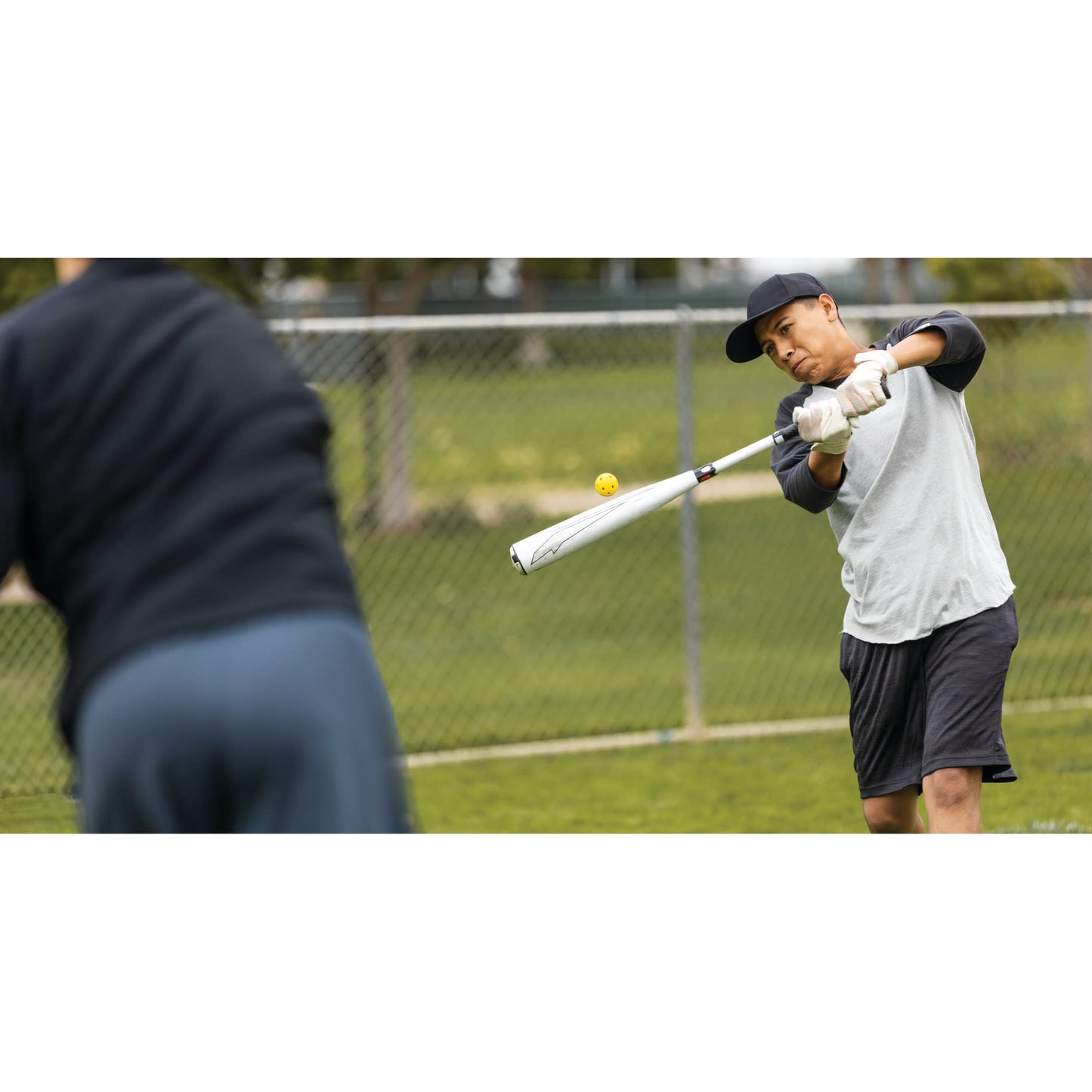 Baseball bat swings at ball during practice on a field outside a chainlink fence