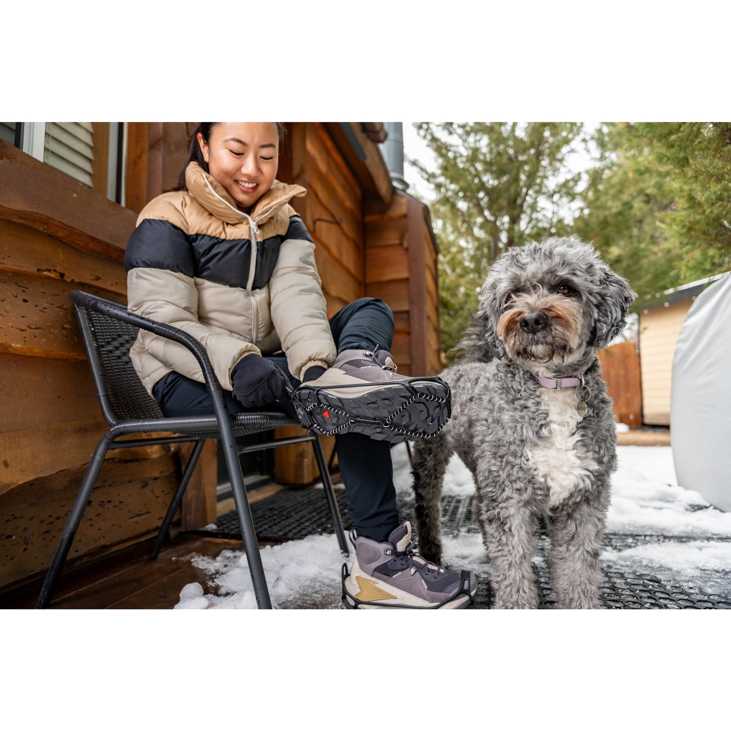 Woman puts on hiking boots outside with dog on snowy porch near cabin