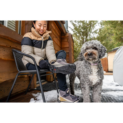 Woman puts on hiking boots outside with dog on snowy porch near cabin