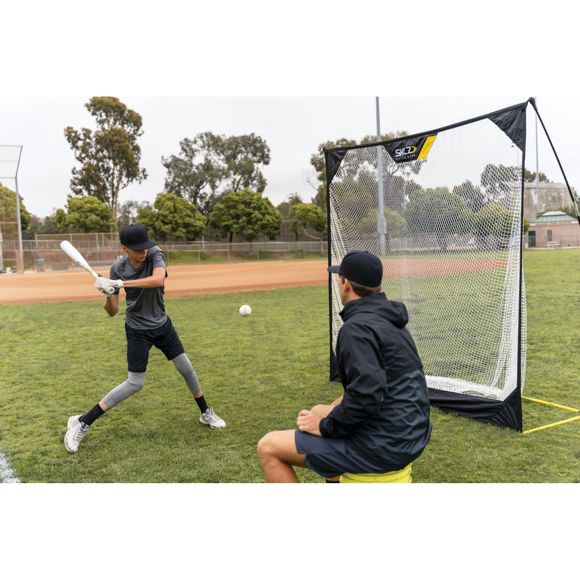 A boy practices batting with a coach nearby during baseball training on a grassy field