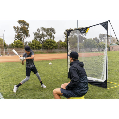 A boy practices batting with a coach nearby during baseball training on a grassy field