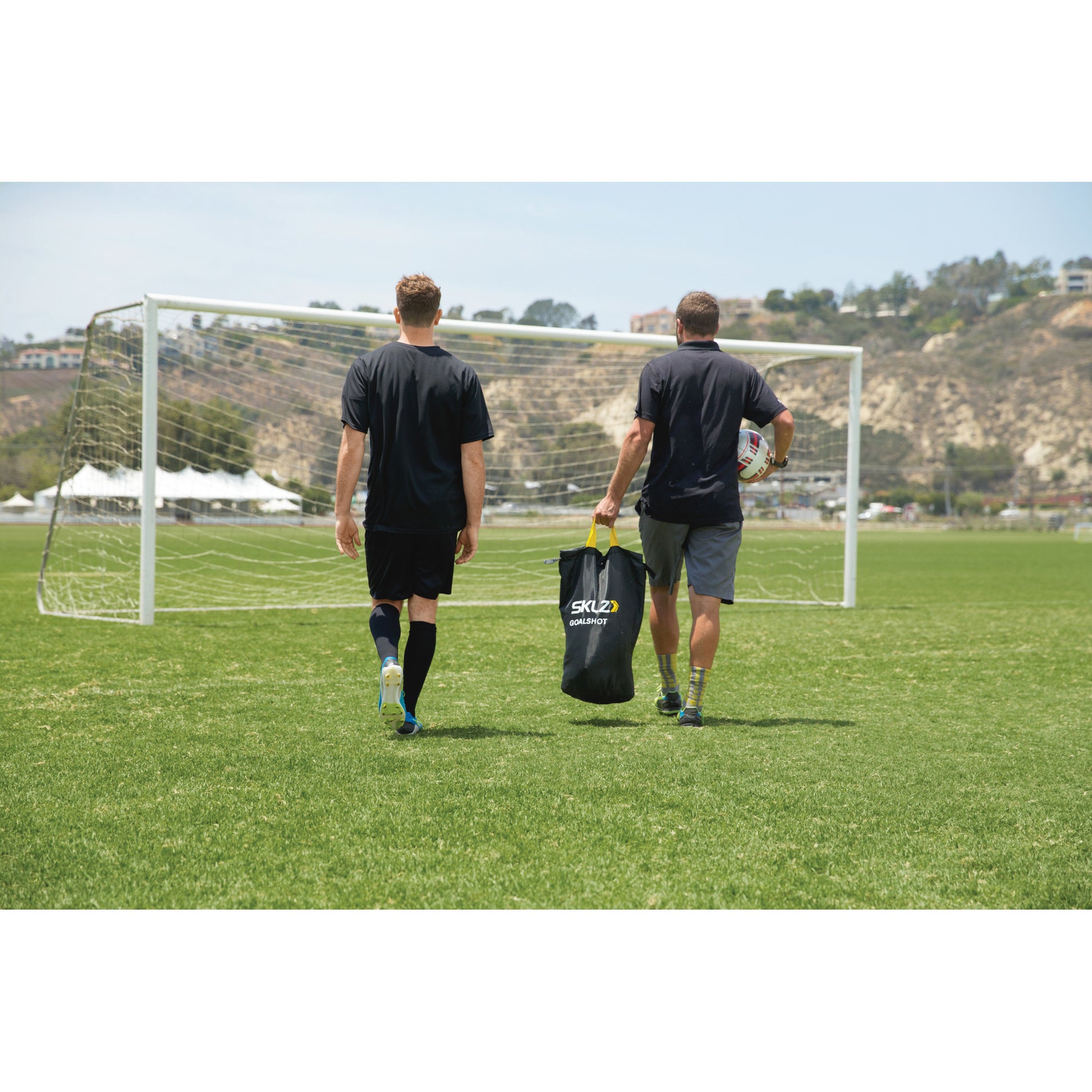 Two players walk across a soccer field carrying gear toward the goal in a training session