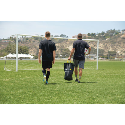 Two players walk across a soccer field carrying gear toward the goal in a training session