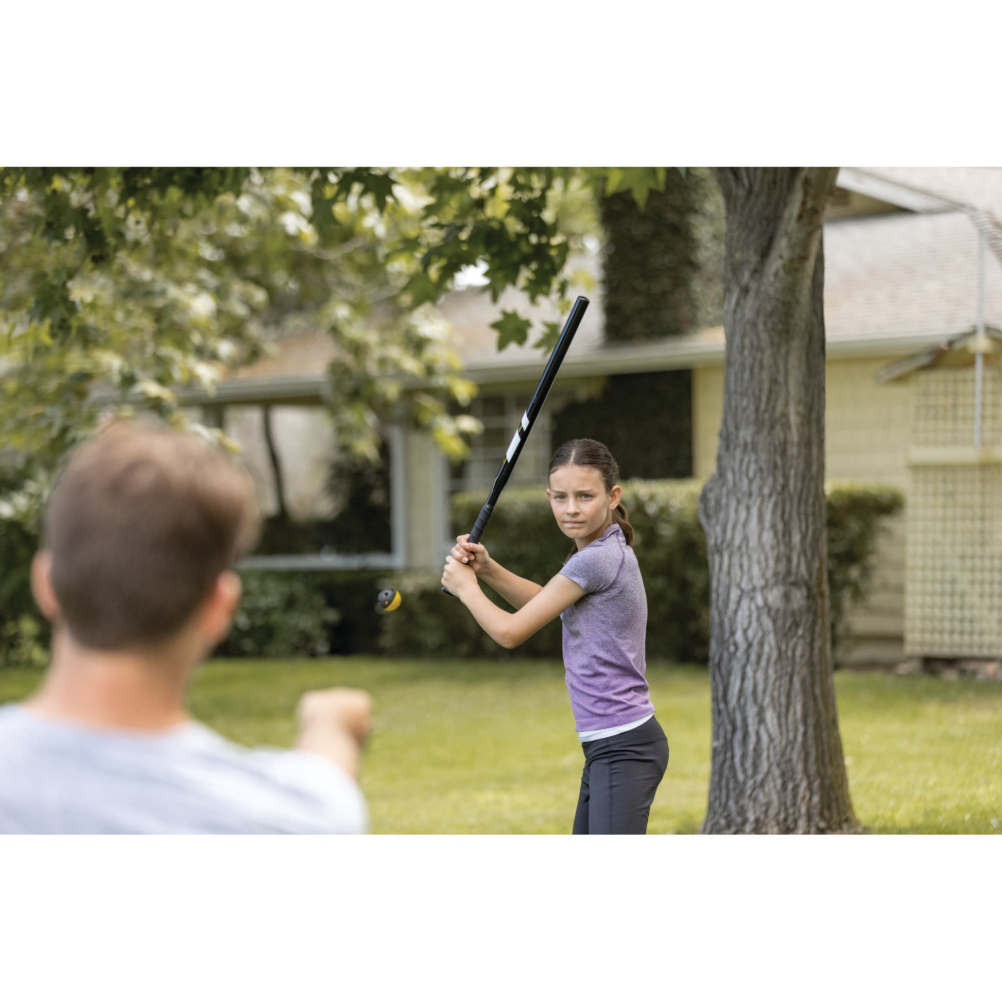 Girl batting at a ball outdoors with a boy in a backyard garden