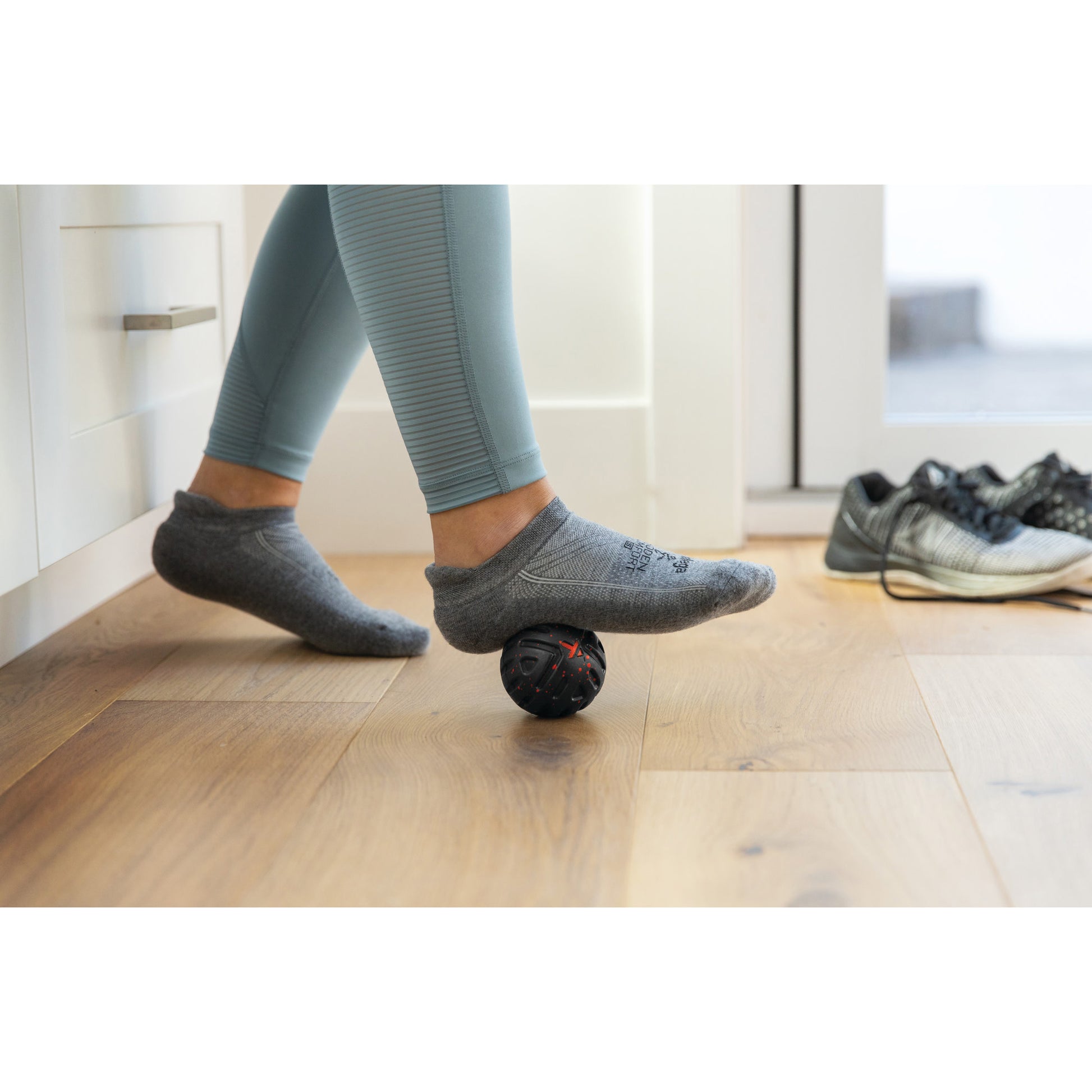 Person's foot rolls over a massage ball on wooden floor in a home environment.