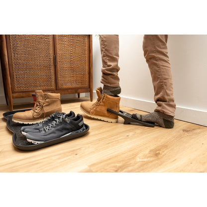 Person using boot jack to remove shoe; sneakers on mat nearby in a room.