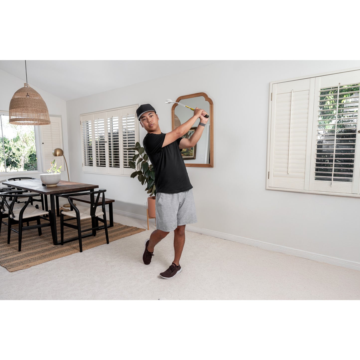 Man practices golf swing indoors in living room with white walls and large windows