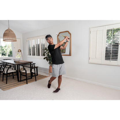 Man practices golf swing indoors in living room with white walls and large windows