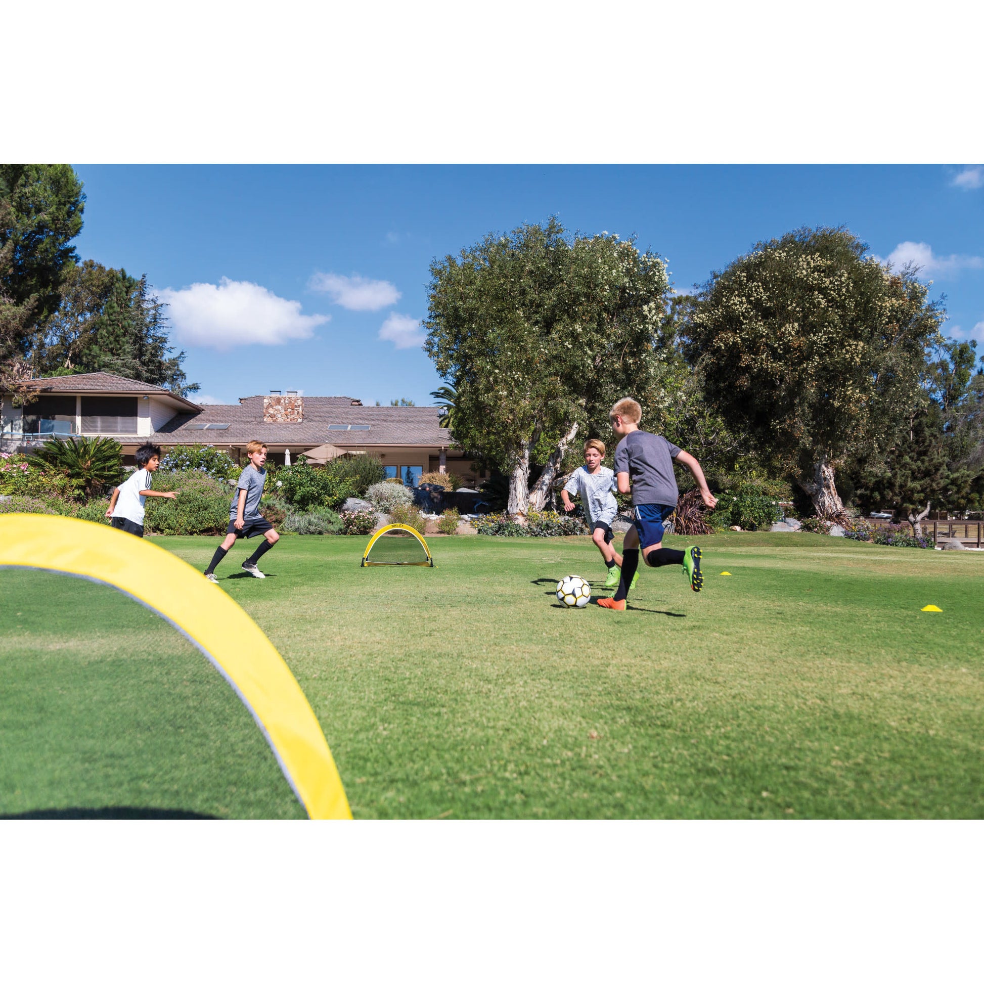 Children play soccer on a grassy field with small hurdles under a blue sky