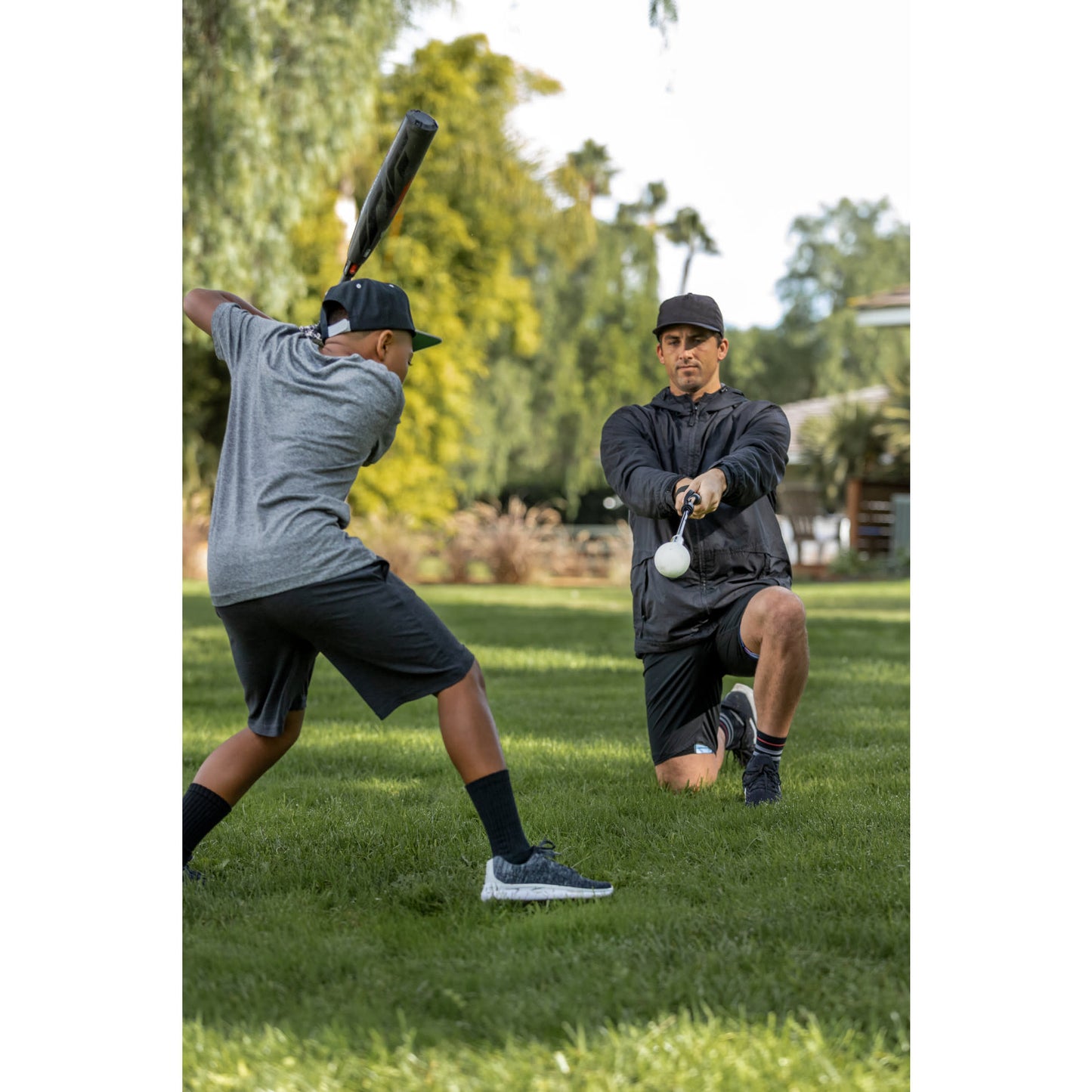 Two boys practice baseball outdoors on a grassy field with trees in the background