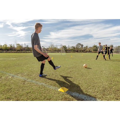 Boy practices soccer dribbling next to yellow cones on a field under a partly cloudy sky