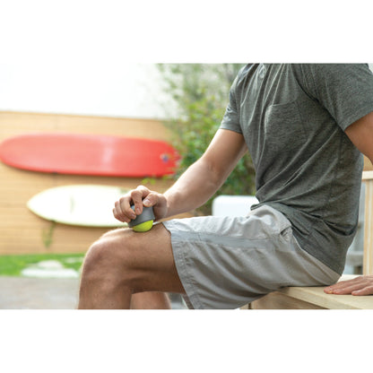Hand holds massage tool on leg outdoors near surfboards in a backyard garden.