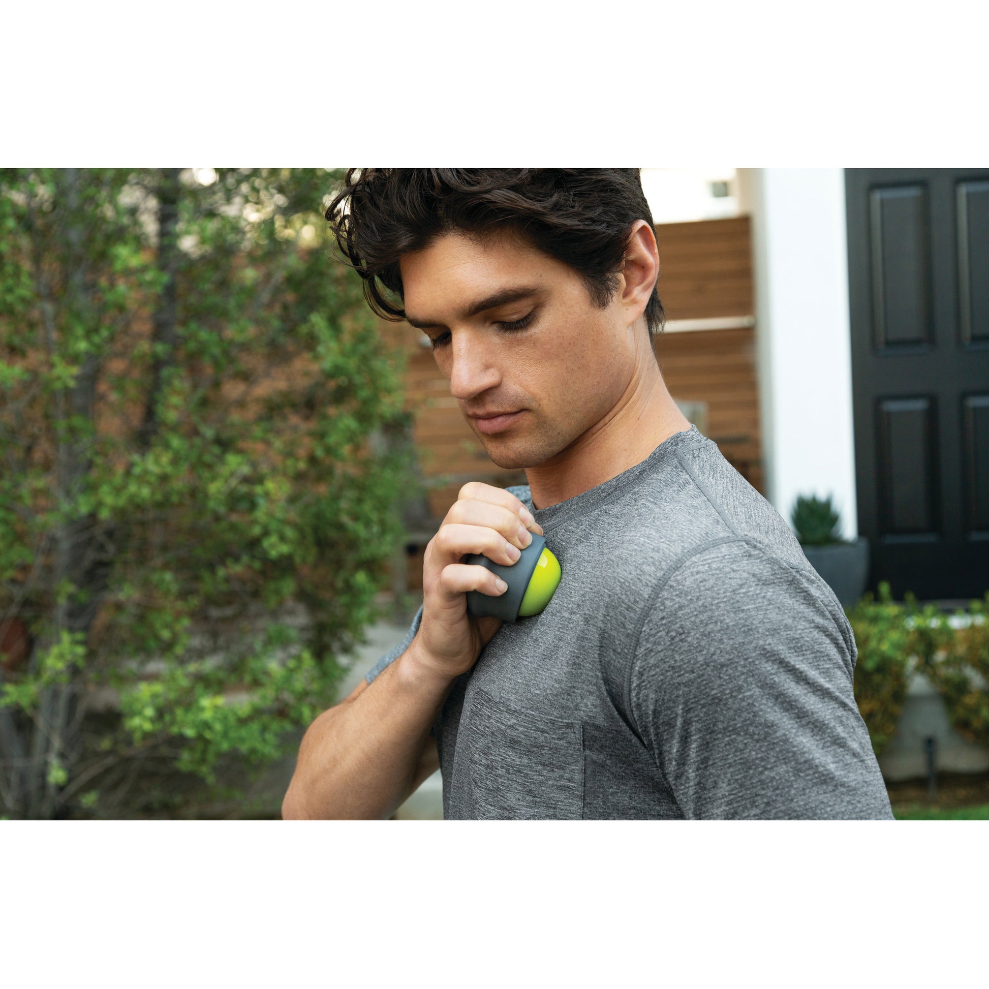 Man presses a portable massager on shoulder outdoors near a house.