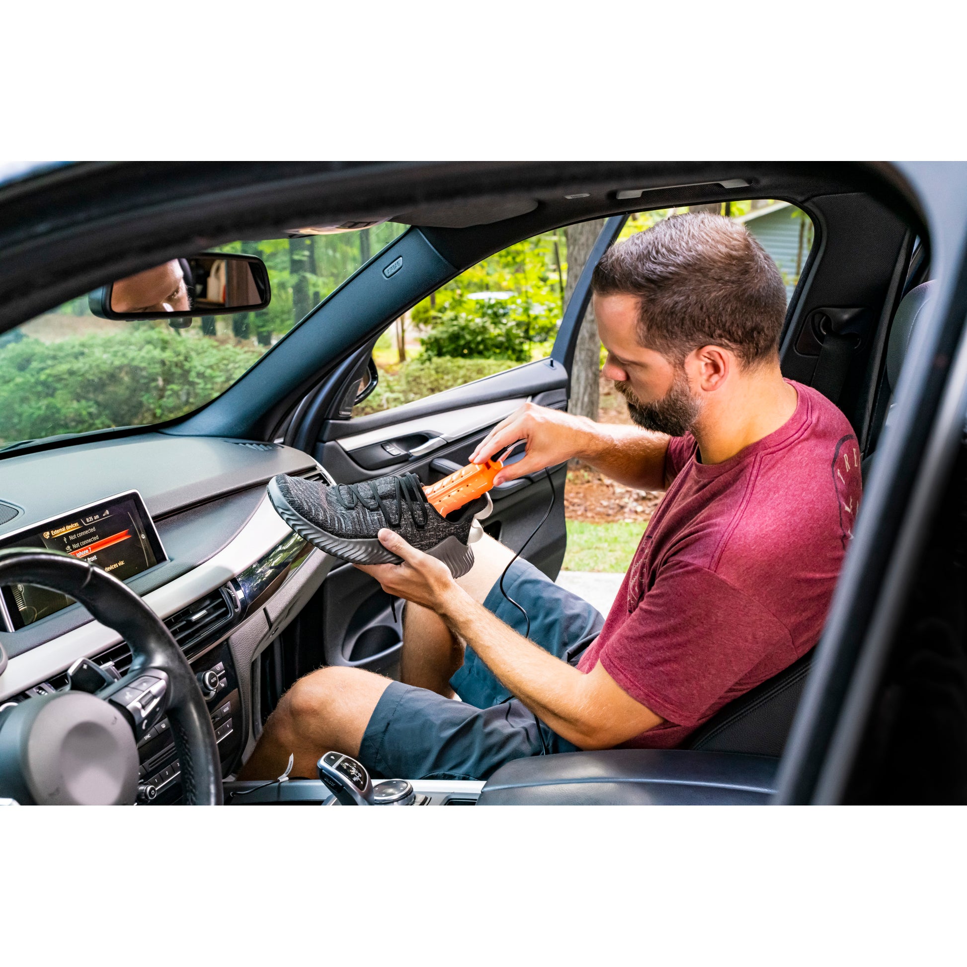 Man inspects a shoe with a shoehorn inside a car on a driveway or street.
