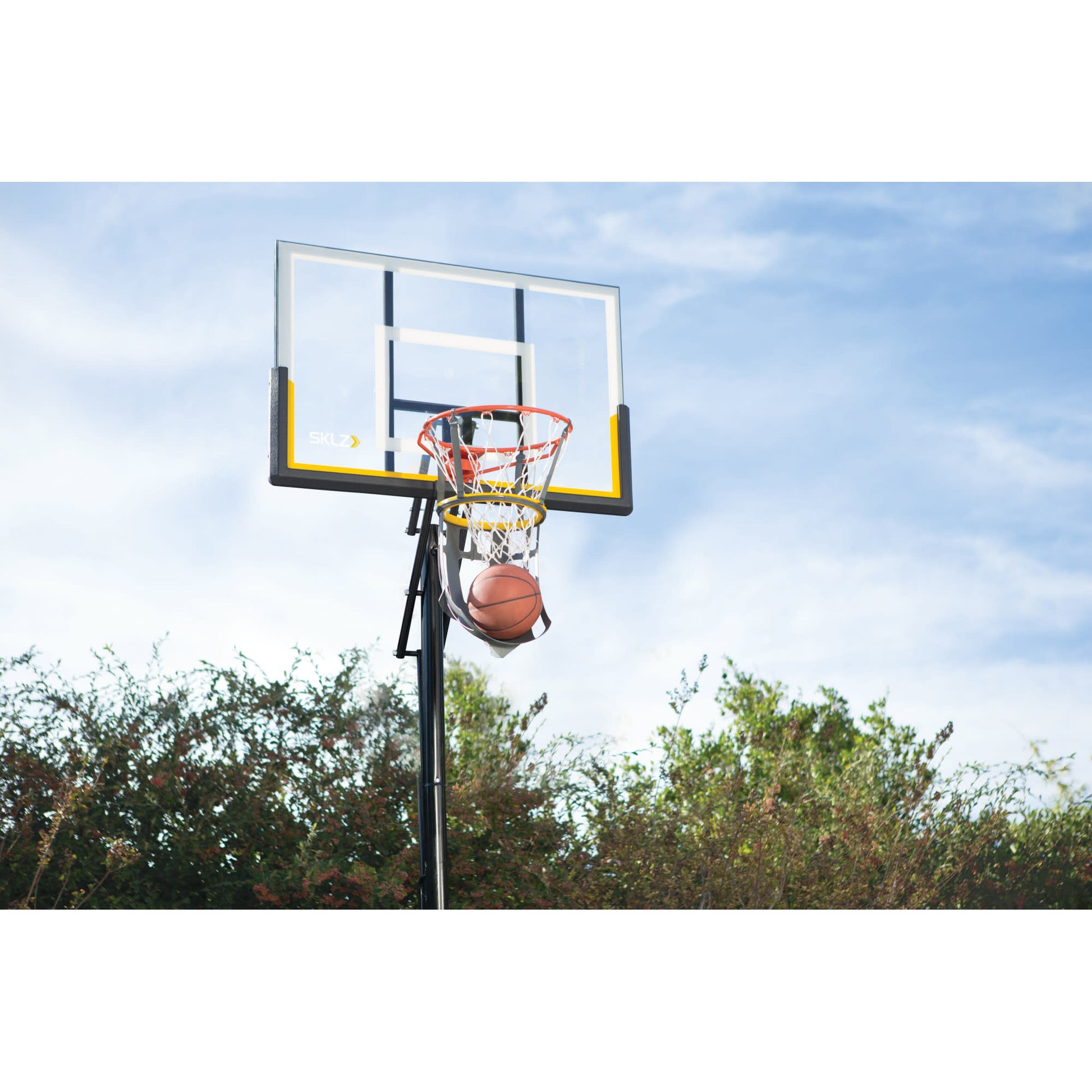 A basketball is in the hoop of a backboard on an outdoor court with trees and sky in the background