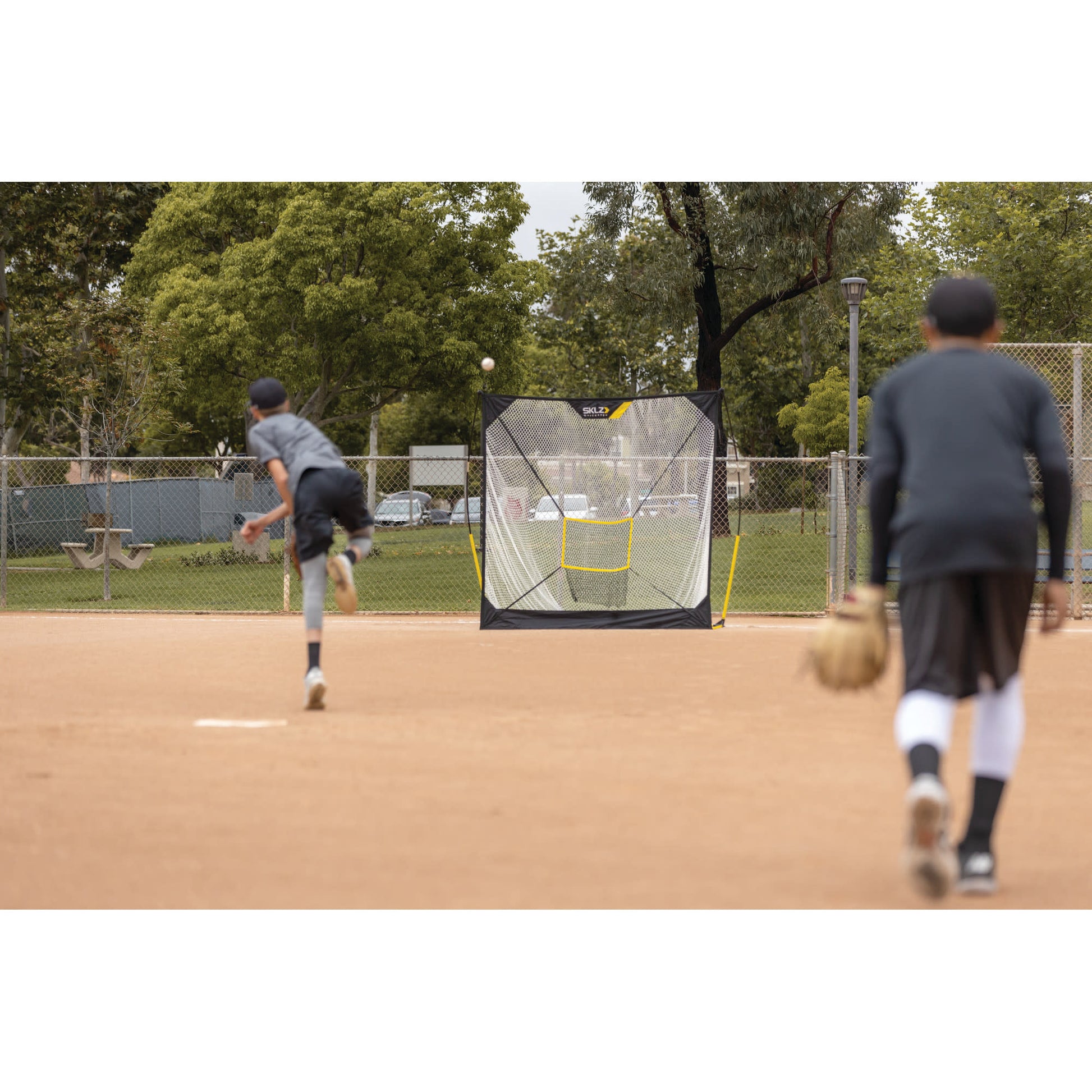 Two boys practice baseball at a park with a pitching screen and trees in the background