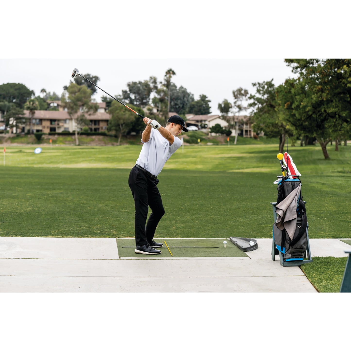Golfer swings club on driving range with golf bag and trees in the background