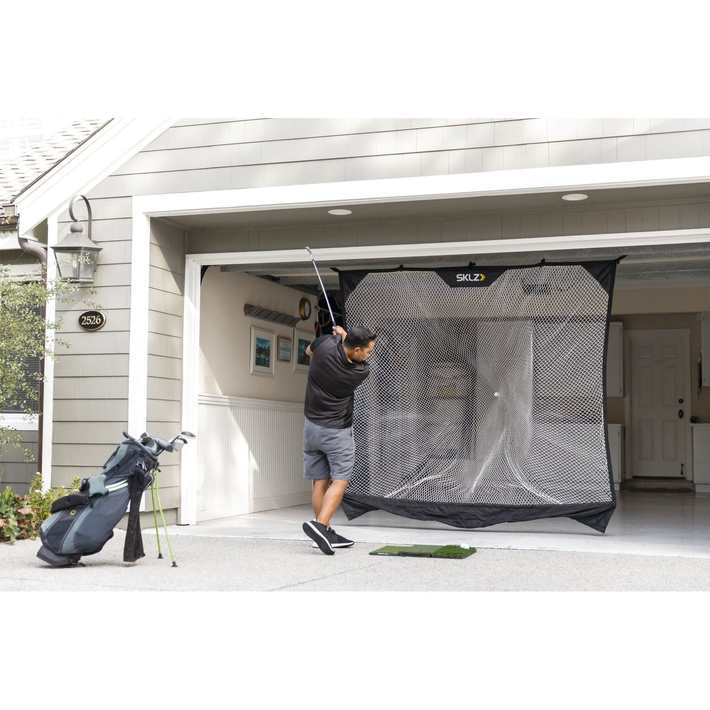 Golfer swings club in garage with golf net and bag nearby during practice