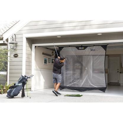Golfer swings club in garage with golf net and bag nearby during practice