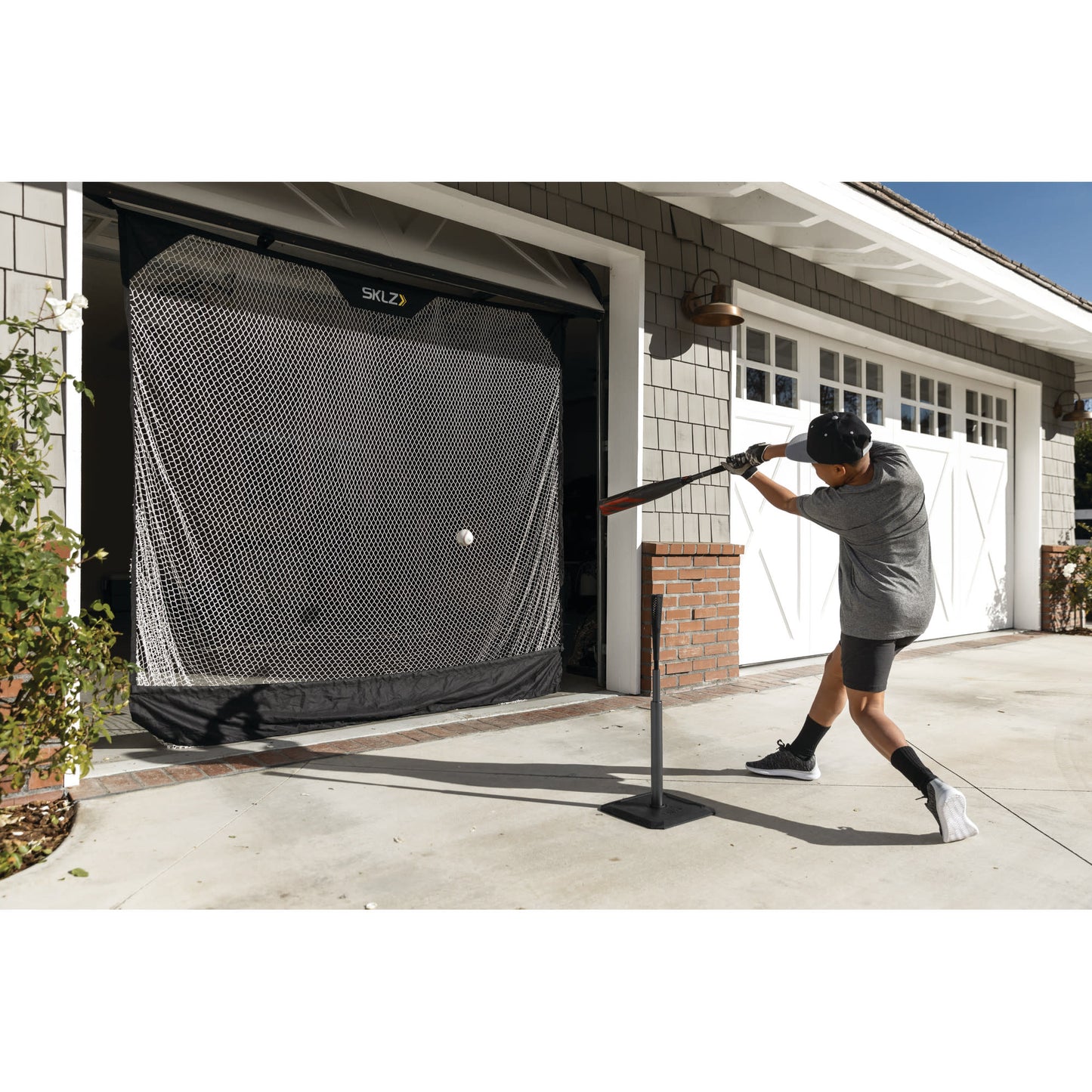 Boy practices baseball hitting against a garage door with a pitching machine outside