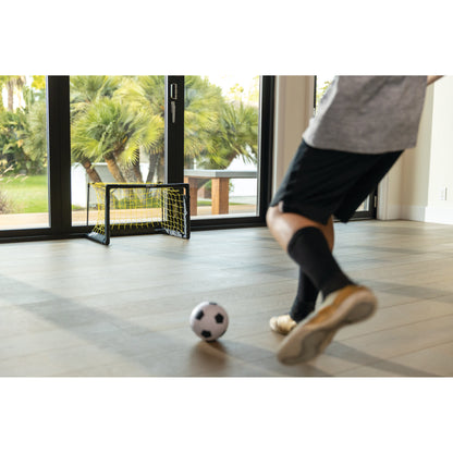 Child kicks a soccer ball toward a small goal indoors near large windows with trees outside