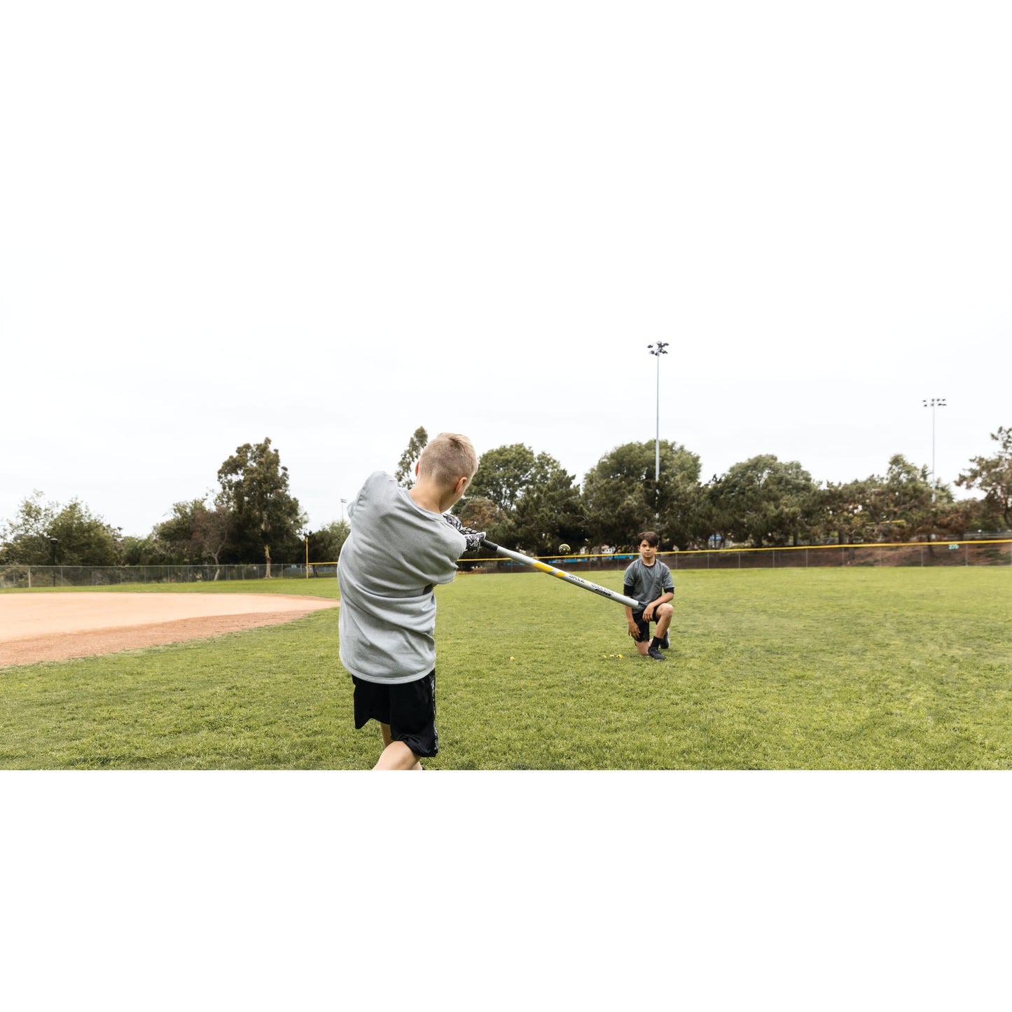 Boy swings a bat at a ball while a man catches the ball on a baseball field