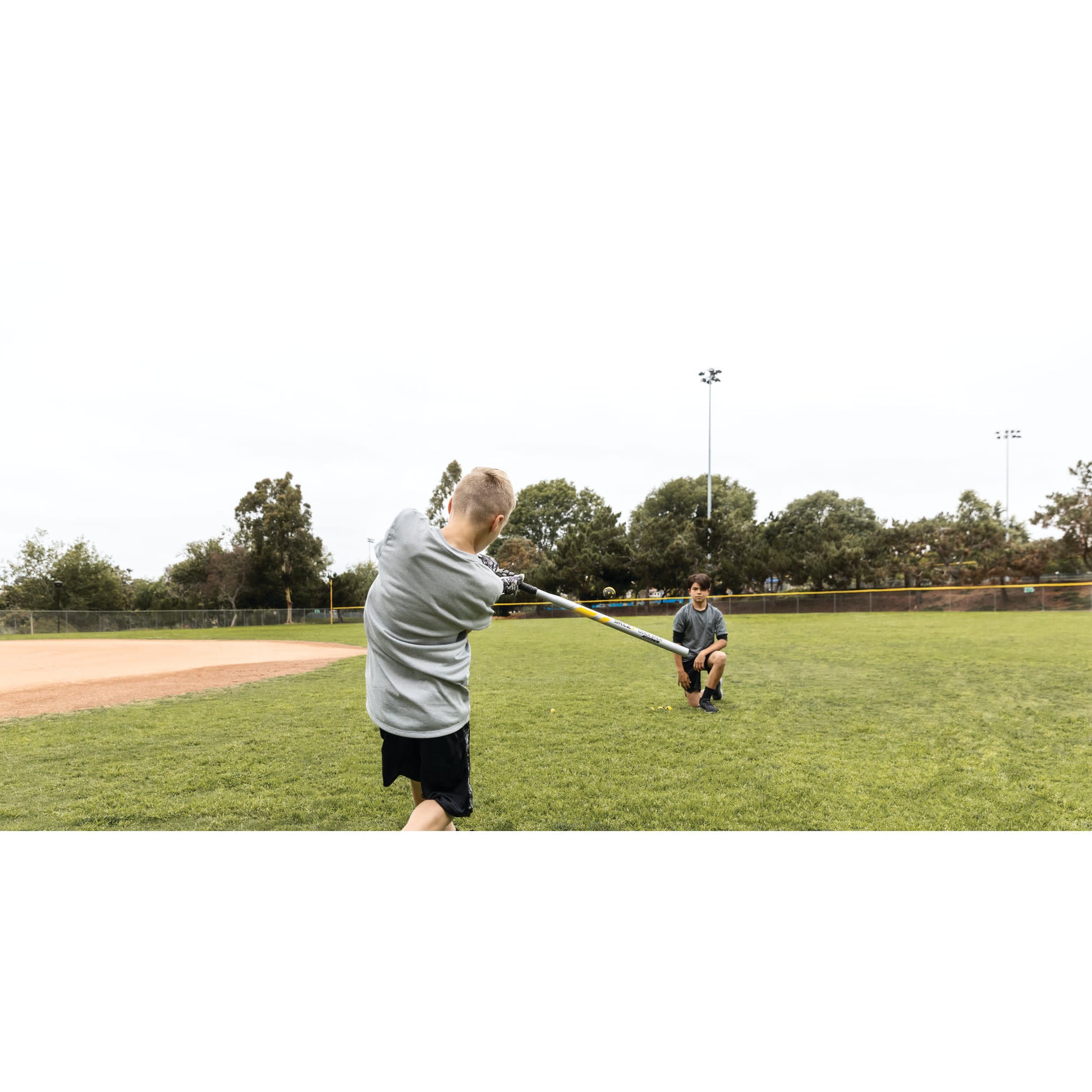 Boy swings a bat at a ball while a man catches the ball on a baseball field