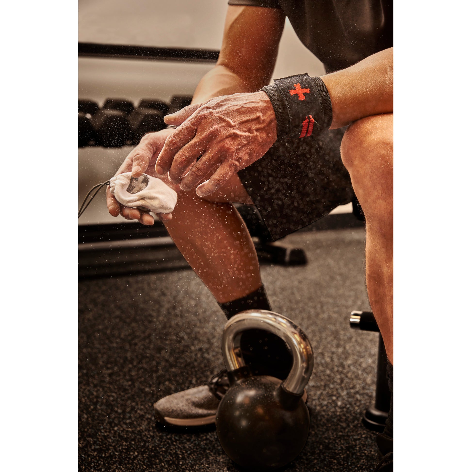 Hands squeezing chalk ball, dust cloud forming, gym setting with kettlebell on floor.