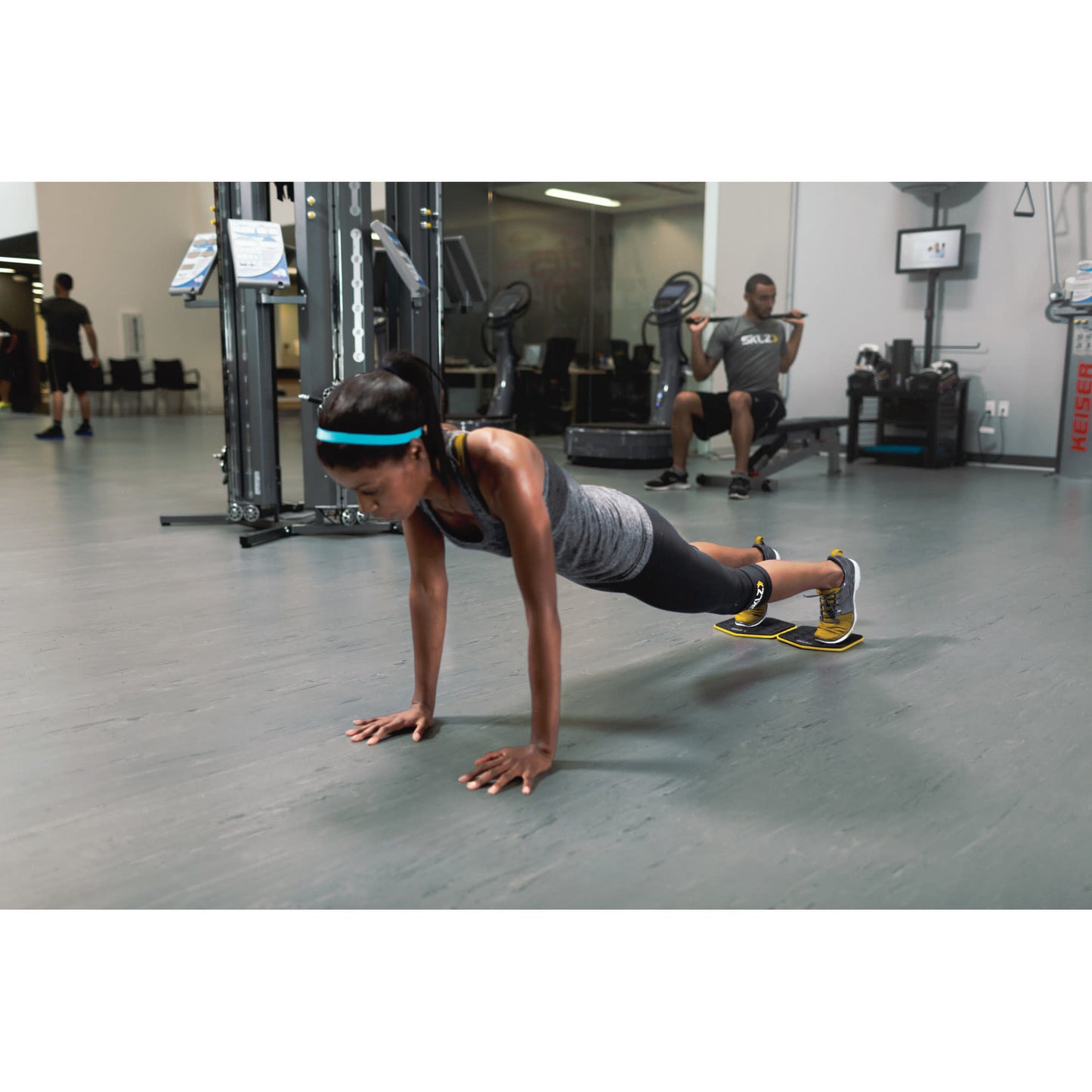Woman performs pushup on mats in gym while man sits on bench in background