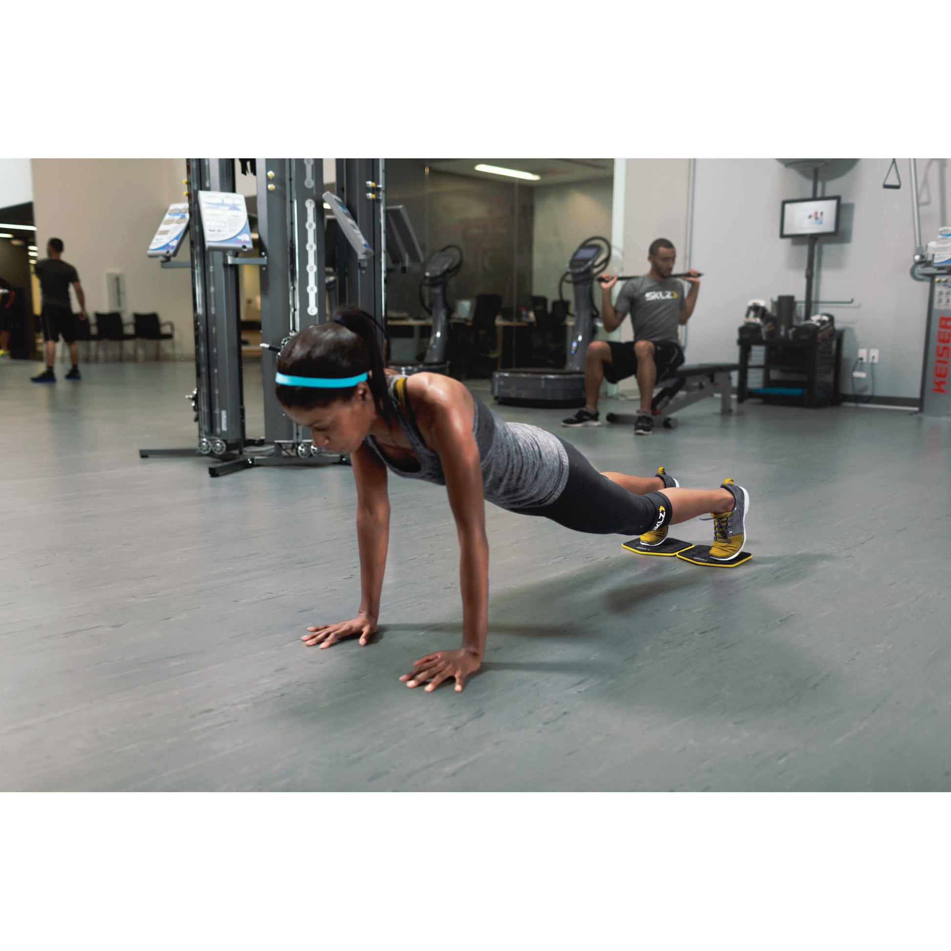 Woman performs pushup on mats in gym while man sits on bench in background