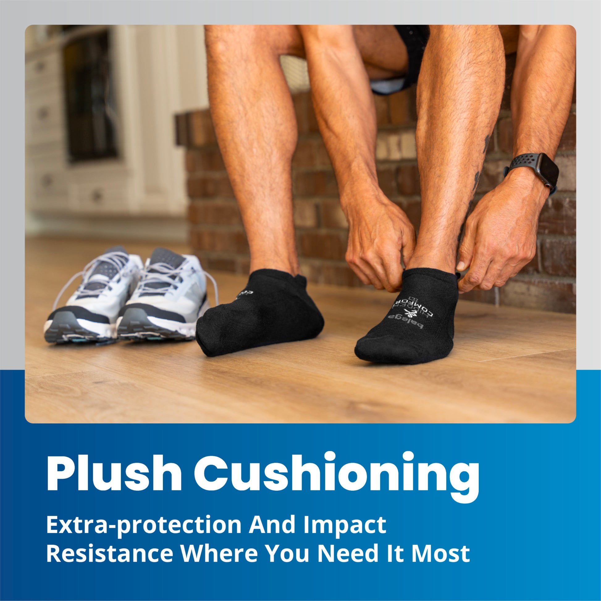 Hands pull black socks on feet; sneakers are on wooden floor by a brick wall.