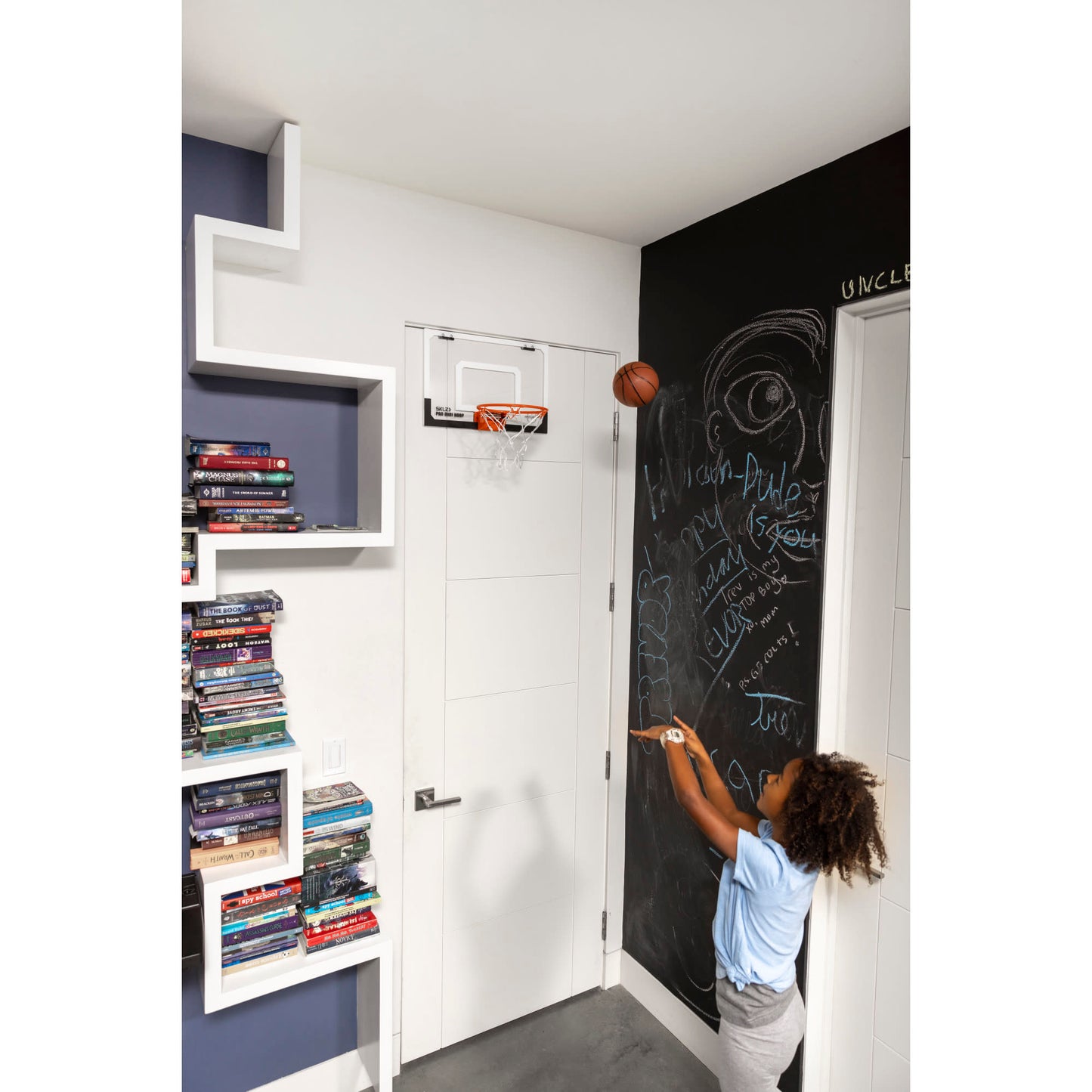 Child writes on blackboard in a room with books and a small basketball hoop