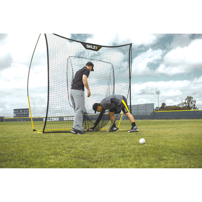 Two players set up a baseball pitching net on a grass field for practice