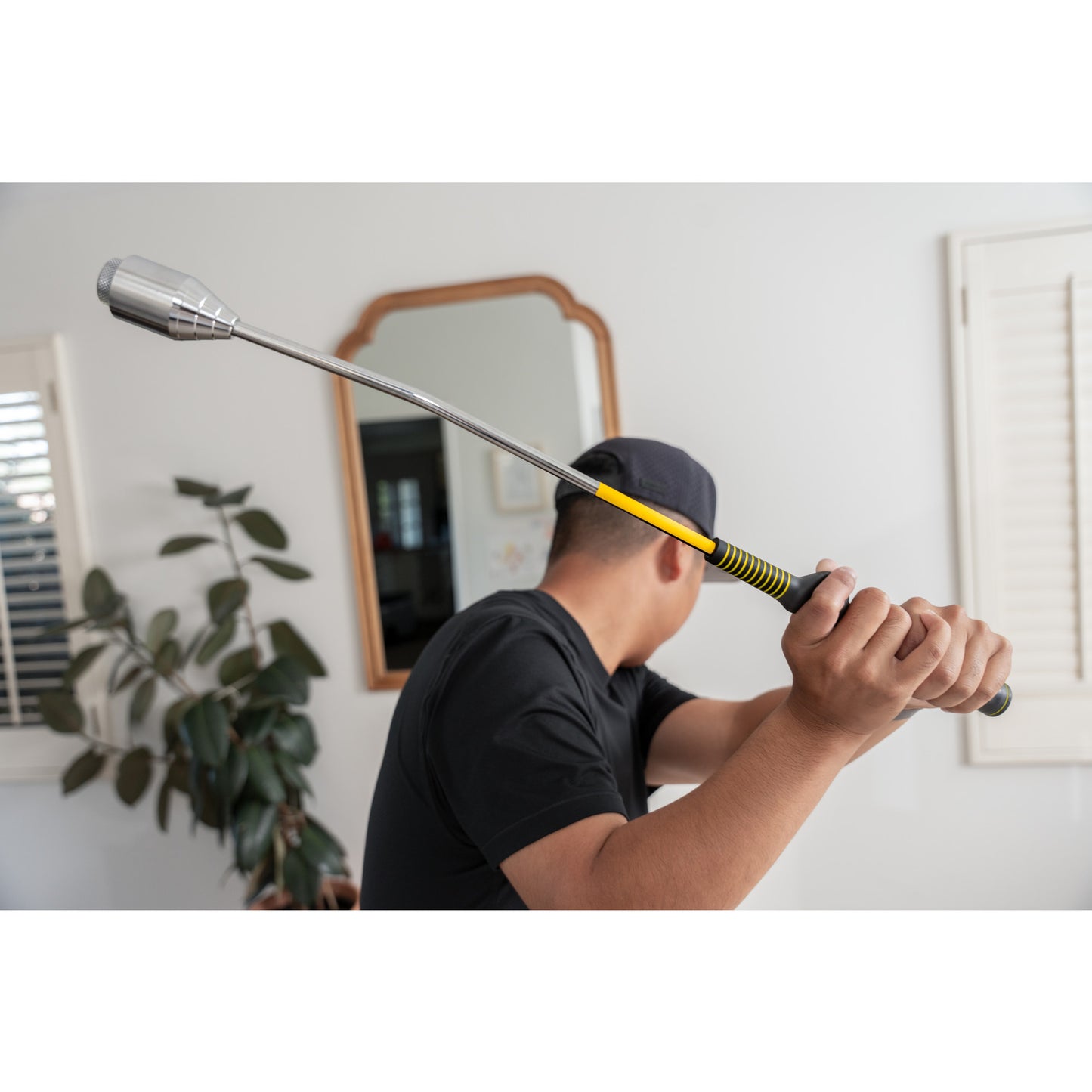 Person aims a golf club in a room with a large mirror window and plant