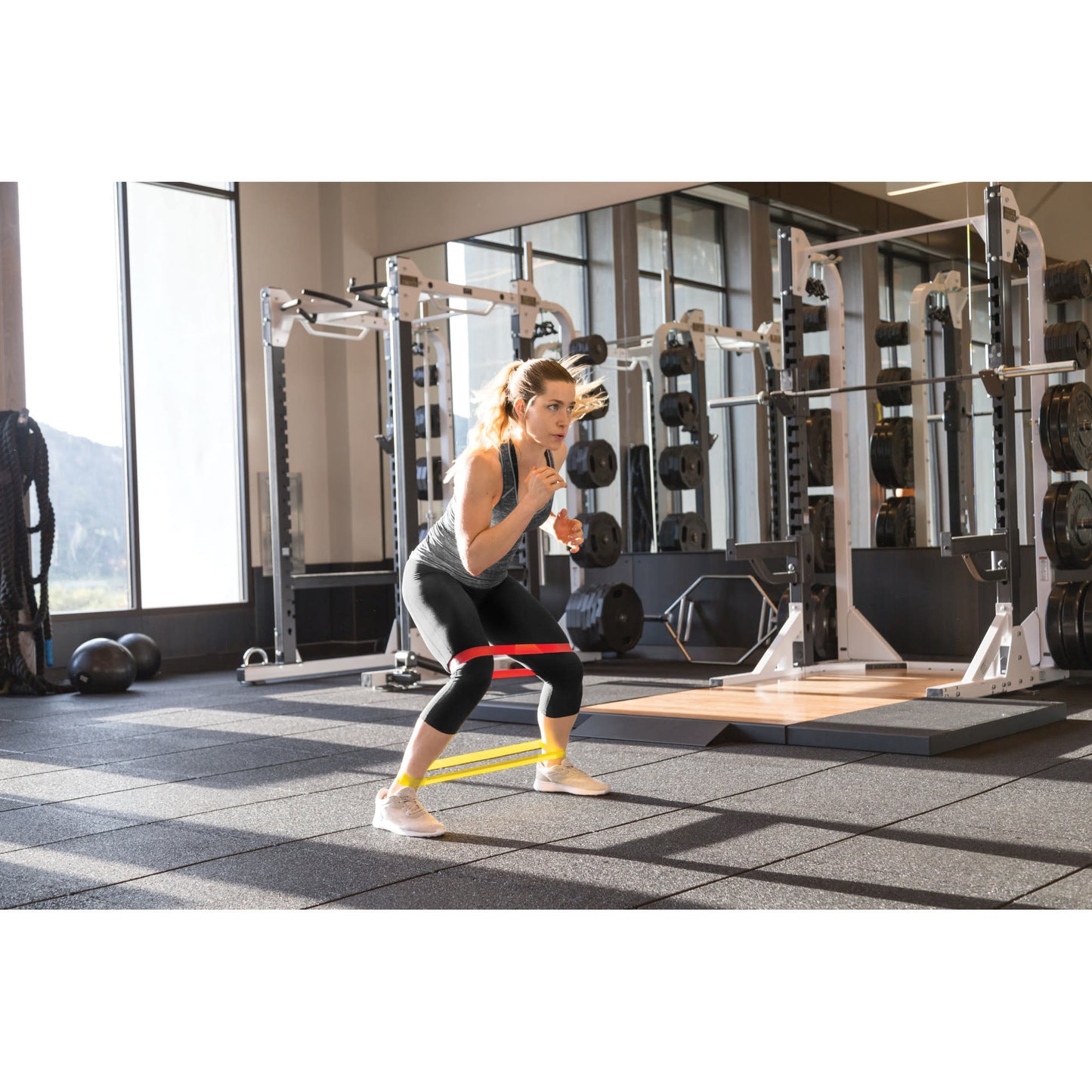 Woman performs resistance band squat in gym with weight training equipment around her