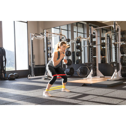 Woman performs resistance band squat in gym with weight training equipment around her