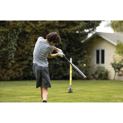 Boy swings a bat at a tennis ball in a backyard with trees and a house in the background