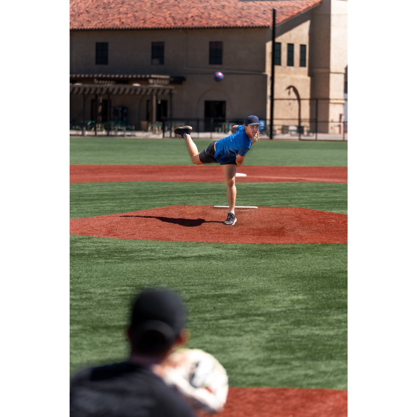 A player pitches a baseball on the mound at a sports field while a fan watches.